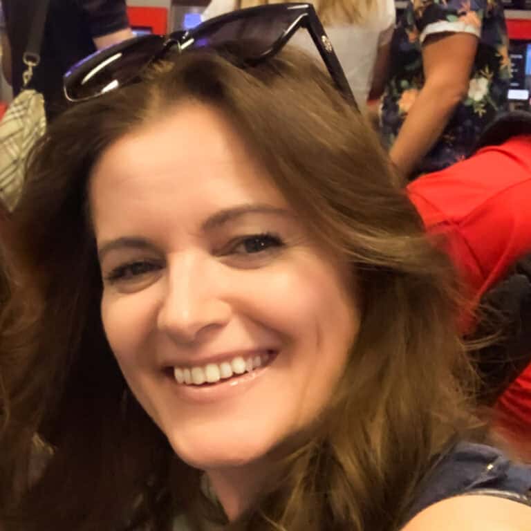 A smiling person with long brown hair, wearing sunglasses on their head, is facing the camera in a crowded indoor space. Other people and indistinct elements of the background can be seen around them.