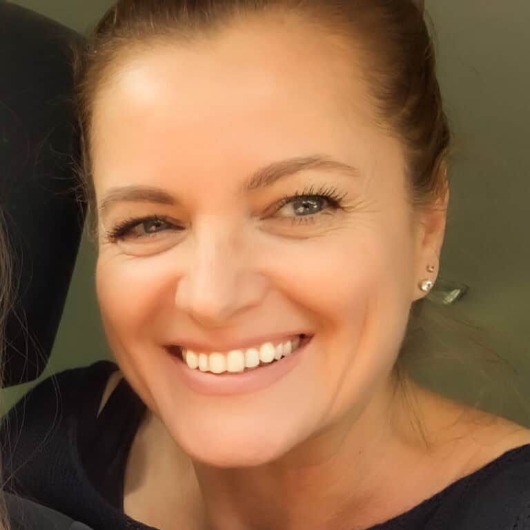 A woman with light skin and brown hair smiles warmly at the camera. She is wearing small, stud earrings and a dark, off-shoulder top. The background is indistinct.