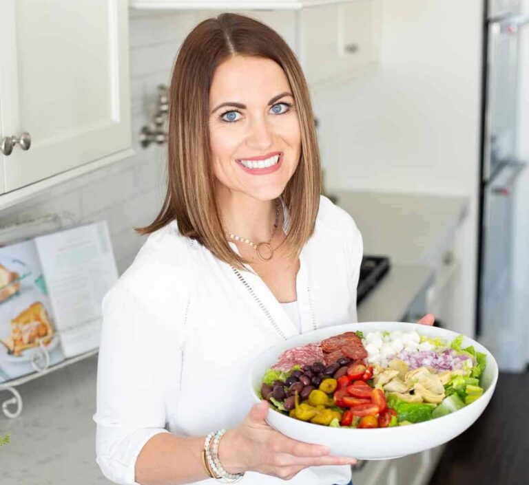 Smiling woman with shoulder-length brown hair holds a large bowl filled with assorted salad ingredients, including cherry tomatoes, olives, onions, and leafy greens. She stands in a bright kitchen, with a cookbook open on the counter behind her.