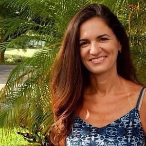 A woman with long brown hair wearing a blue patterned sleeveless top smiles while standing outdoors in front of green palm leaves.