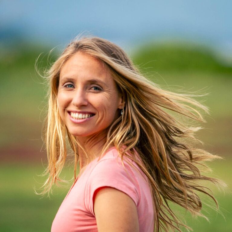 Smiling woman with long blonde hair wears a pink shirt, standing outdoors with green fields and a blue sky in the background. Her hair is gently blowing in the wind.