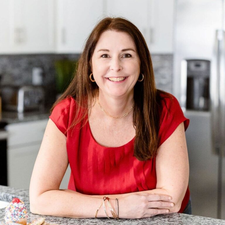 A woman with long brown hair, wearing a red short-sleeve shirt and gold jewelry, is smiling while leaning on a kitchen counter. The background shows a modern kitchen with white cabinets and stainless steel appliances. A decorated cupcake is visible on the counter.