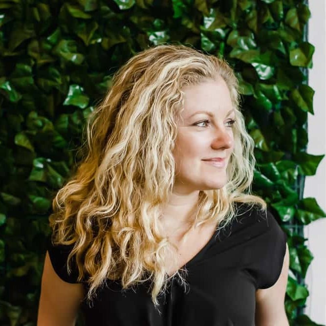 A person with wavy blonde hair is wearing a black top while standing in front of a background filled with green leaves. They are looking to the side and smiling.