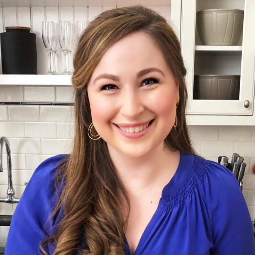 A person with long brown hair smiles at the camera in a kitchen setting. They are wearing a blue top and gold hoop earrings. Shelves with dishes and glasses are visible in the background.