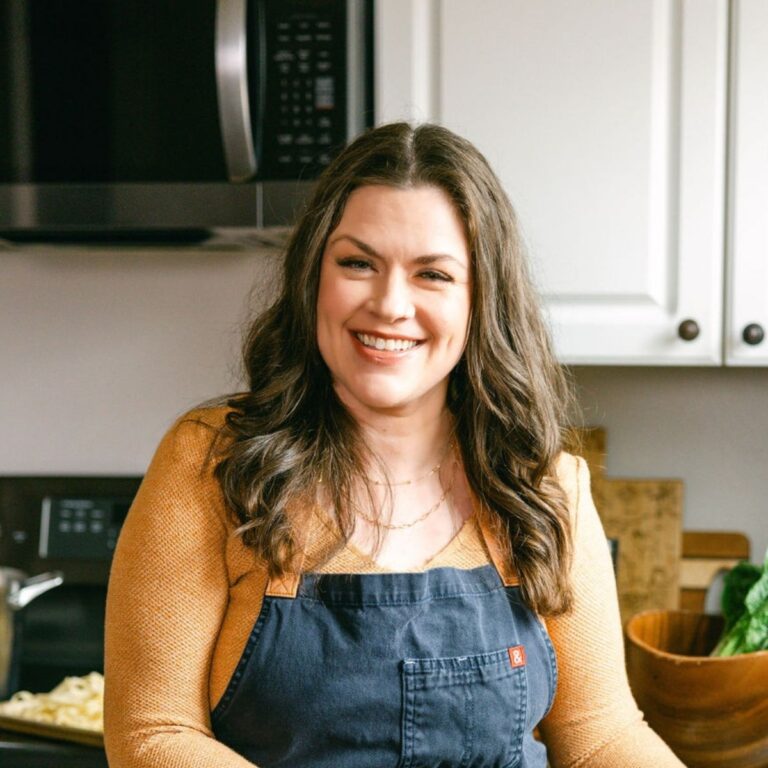 Smiling woman with long brown hair, wearing a mustard yellow top and a dark apron, stands in a kitchen. The background shows white cabinets, a microwave, and a partial view of a wooden bowl with leafy greens.
