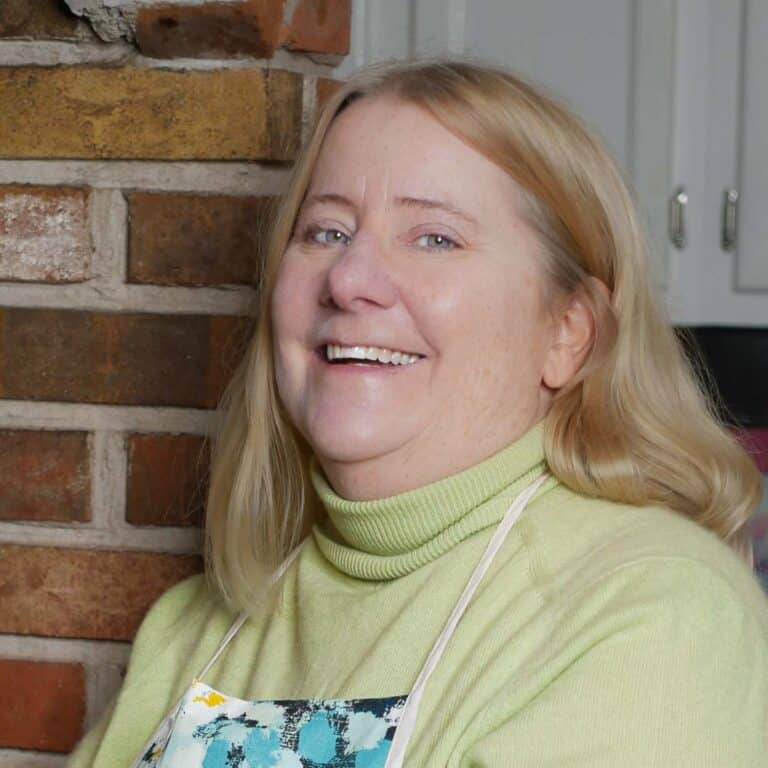 A woman with blonde hair and a cheerful expression leans against a brick wall. She is wearing a lime green turtleneck sweater and a patterned apron. The background shows a portion of a kitchen with white cabinets.