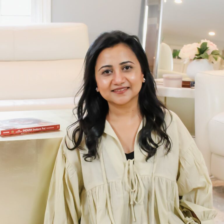 A person with long dark hair is seated in a light-colored room, wearing a beige blouse. A book titled "Indian Instant Pot" is on the table beside them. They are smiling slightly, and there are white flowers in the background.