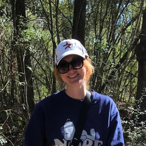 A person wearing a baseball cap and sunglasses smiles while standing in a sunlit forest. They are dressed in a dark sweatshirt with a bag strap across their chest, surrounded by trees and greenery.