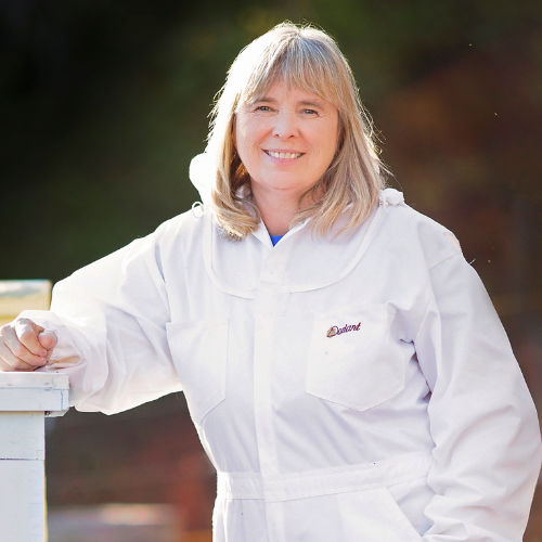 A person wearing a white beekeeping suit stands outdoors, smiling. They lean on a white surface with a relaxed posture. The background is softly blurred, suggesting a natural setting.