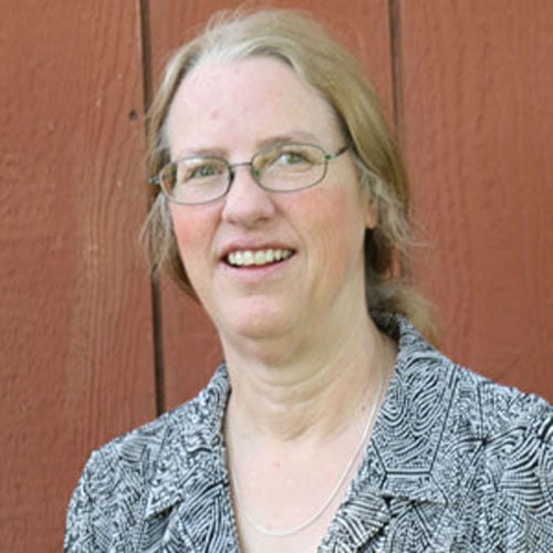 A woman with light hair, glasses, and a patterned black-and-white top stands in front of a reddish-brown wooden wall. She is smiling and looking slightly off camera. Her hair is pulled back, and she is wearing a necklace.