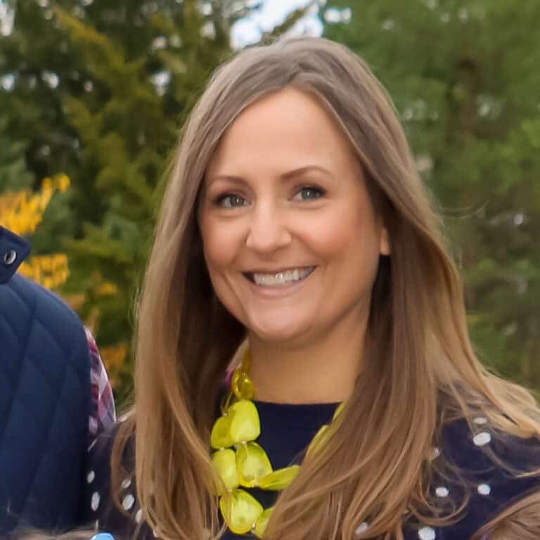 A woman with long blonde hair and a big smile stands outdoors wearing a navy blue polka-dot top and a bright yellow necklace. The background features green foliage and trees.