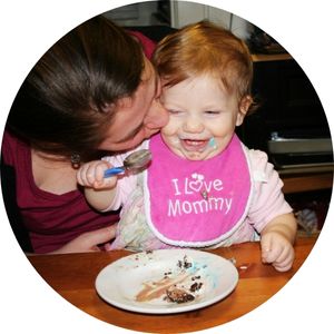 A woman lovingly kisses a smiling toddler who is wearing a pink bib that says "I ♥ Mommy." The toddler is holding a spoon and has food smeared around their mouth, while an empty plate with some remnants of food is on the table in front of them.