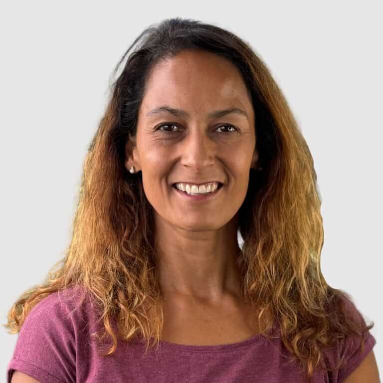 A woman with shoulder-length wavy hair, blending shades of brown and blonde, smiles warmly. She is wearing a maroon top and small earrings. The background is plain and light-colored.