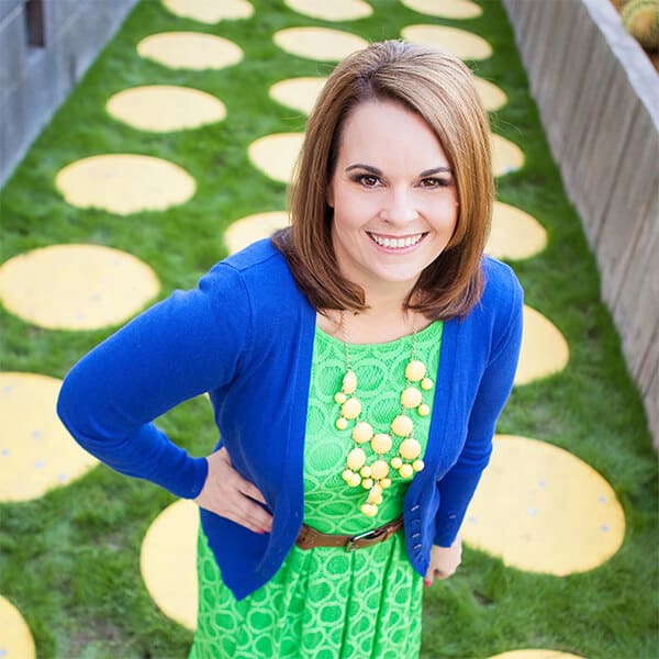A smiling woman in a green dress and blue sweater stands on grass decorated with large yellow circles, looking up at the camera. She wears a chunky yellow necklace and has shoulder-length brown hair.