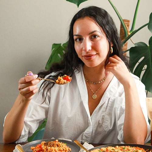 A person with long dark hair is sitting at a table, holding a spoonful of food. They are wearing a white shirt and jewelry. Plates of food are on the table, and there are green plants in the background.