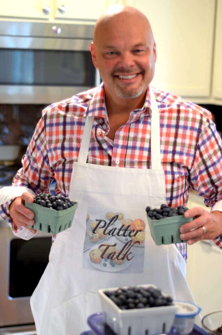 A man in a checkered shirt and white apron stands in a kitchen, smiling and holding containers filled with blueberries. The apron has a "Platter Talk" logo on it. A tray with additional blueberries is on the counter in front of him.