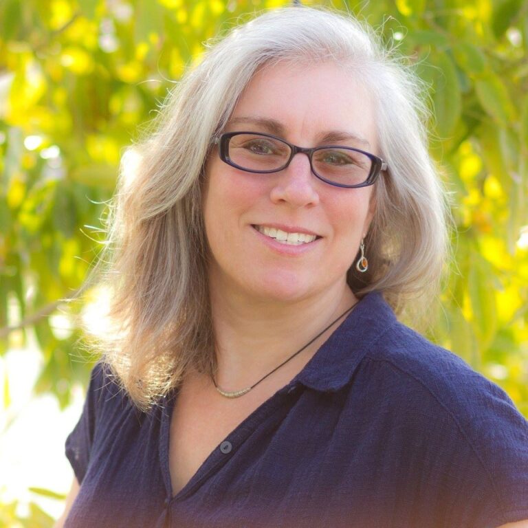A person with shoulder-length gray hair, wearing glasses and a navy blue top, is smiling. They are standing outside in front of a backdrop of green leaves and sunlight.