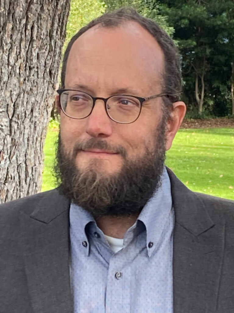 A man with a beard and glasses is standing outdoors near a tree. He is wearing a gray suit jacket over a light blue shirt. The background features green grass and trees.