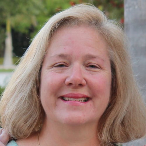 A woman with shoulder-length blonde hair smiles at the camera. She is outside, and there are green trees in the blurred background.