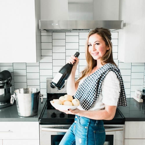 A woman in a kitchen holds a bottle of wine and a plate of rolls. She wears a checkered towel over her shoulder and stands in front of a modern stove with a tile backsplash. An ice bucket and a mixer are on the counter.