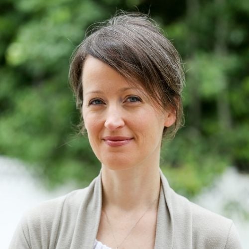 A person with short brown hair smiles slightly while standing outdoors. They wear a light beige cardigan over a white top. The background is blurred greenery.