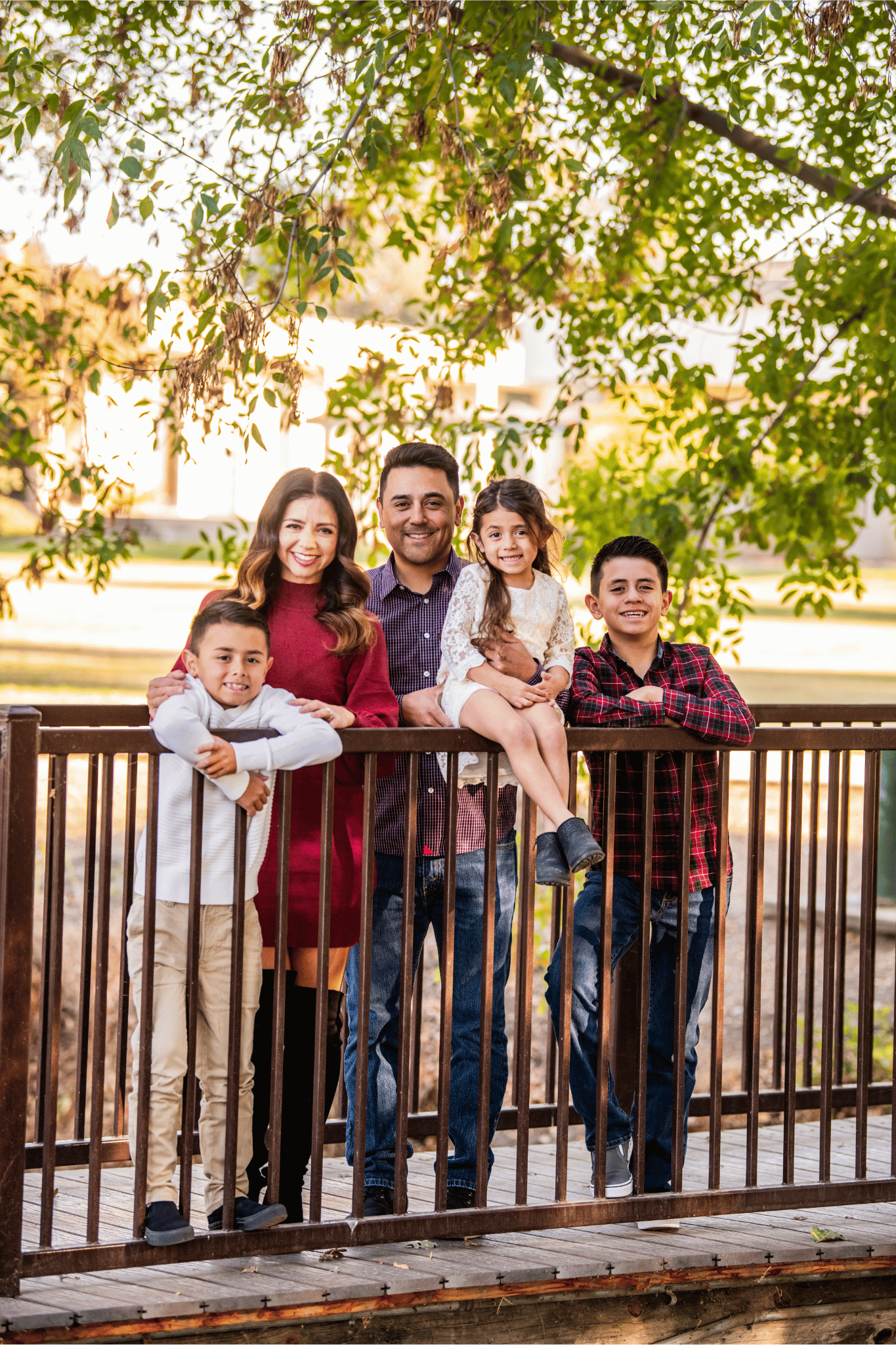 A family of five poses together on a wooden bridge surrounded by greenery. Two boys stand on either side of the parents, while a young girl sits on the railing. The scene is bright and cheerful, suggesting a warm day.
