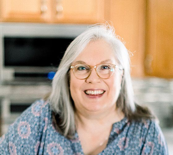 A woman with long gray hair and glasses is smiling in a warmly lit kitchen. She is wearing a blue patterned top and appears relaxed and happy. The background shows wooden cabinets and stainless steel appliances slightly out of focus.