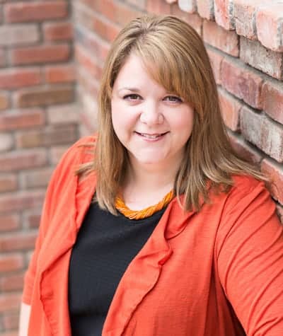 A woman with shoulder-length light brown hair is smiling at the camera. She is wearing an orange blazer over a black top and a beaded necklace in front of a brick wall background.