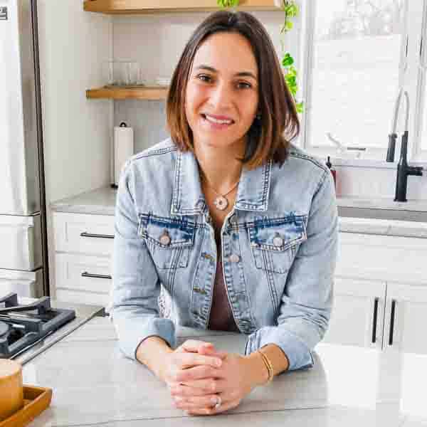 A person with shoulder-length brown hair, wearing a light denim jacket, smiles while leaning on a white kitchen counter. There are green plants and a sink with a black faucet in the background.