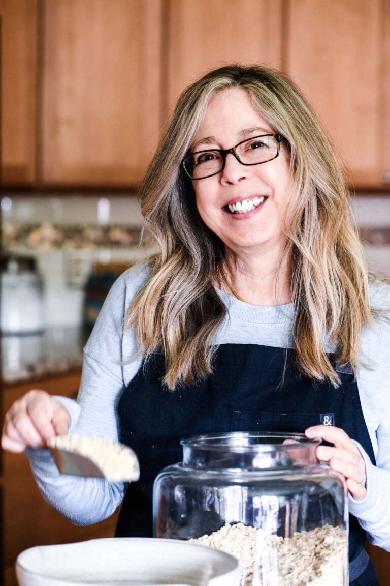 A woman with long hair and glasses smiles while scooping oats from a large jar in a kitchen. She wears a light blue sweater and a black apron. The background features wooden cabinets and kitchen items.