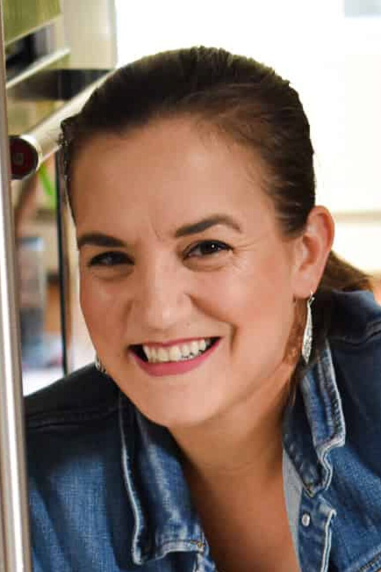 Smiling woman with dark hair pulled back, wearing a denim jacket and hoop earrings. She is positioned near a metallic surface, possibly a kitchen setting. The background is softly blurred.