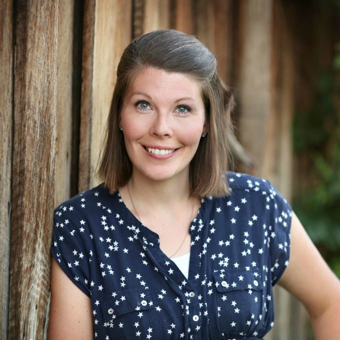A woman with shoulder-length hair smiles while leaning against a wooden wall. She is wearing a blue blouse with white stars and has one hand on her hip. The background is a rustic wooden surface.