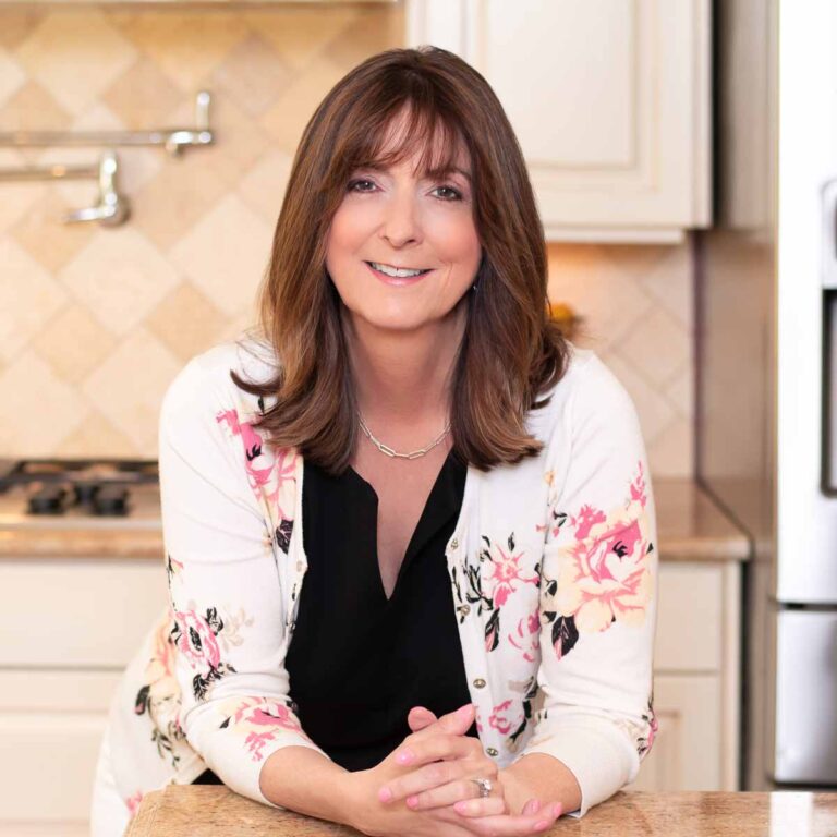 A woman with shoulder-length brown hair and bangs is standing in a kitchen with beige tile backsplash and light-colored cabinets. She is wearing a white cardigan with pink floral patterns over a black top and smiling at the camera.
