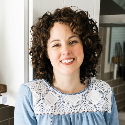 A person with curly hair and a friendly smile is standing in a kitchen. They are wearing a blue top with white lace detailing. The background shows kitchen utensils and part of a cabinet.