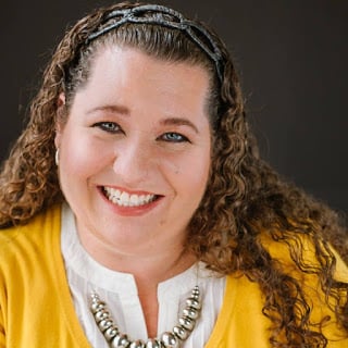 Smiling person with long curly hair wearing a yellow cardigan over a white blouse and a beaded necklace, posed against a dark background.