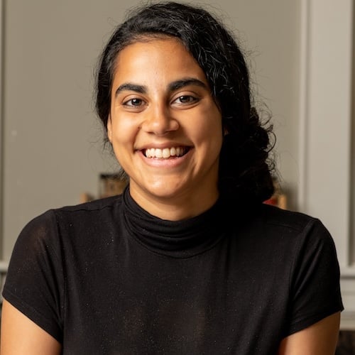 A person with long, dark hair is smiling at the camera. They are wearing a black shirt and are positioned in front of a neutral background with soft lighting.