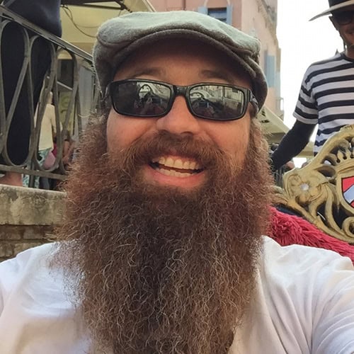 A bearded man wearing sunglasses, a gray flat cap, and a white t-shirt is smiling at the camera. He is standing outdoors near a railing, with a bit of another person visible in the background.