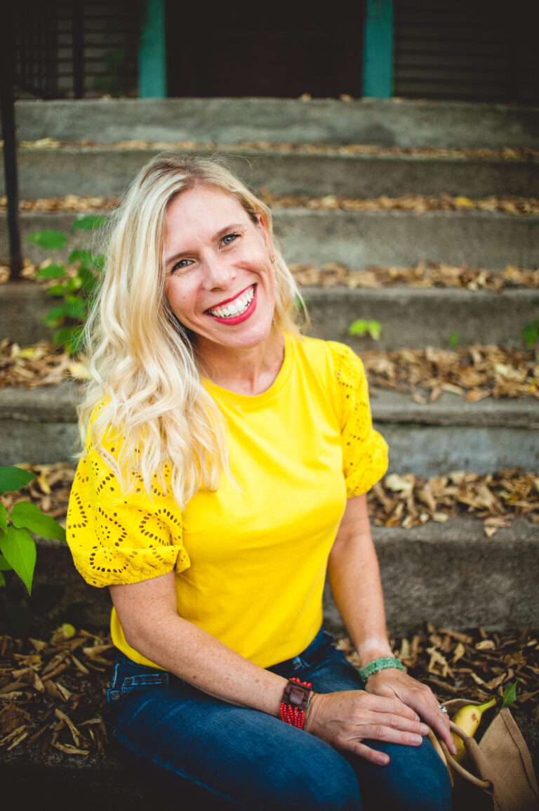 A woman with long, wavy blonde hair and a bright smile sits outdoors on stone steps. She is wearing a yellow top with lacy short sleeves and blue jeans. The background includes green foliage and dried leaves scattered on the steps.