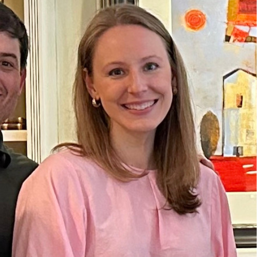 A woman with long, straight hair is smiling at the camera. She is wearing a pink top and gold hoop earrings. Behind her, there is an abstract painting with orange and red tones, depicting geometric shapes.