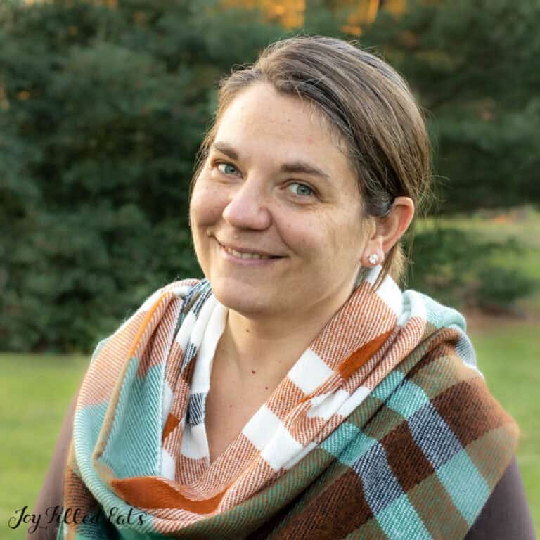 A woman with light skin and brown hair pulled back smiles at the camera. She is wearing a colorful plaid scarf over a dark top. The background is an outdoor setting with lush greenery.