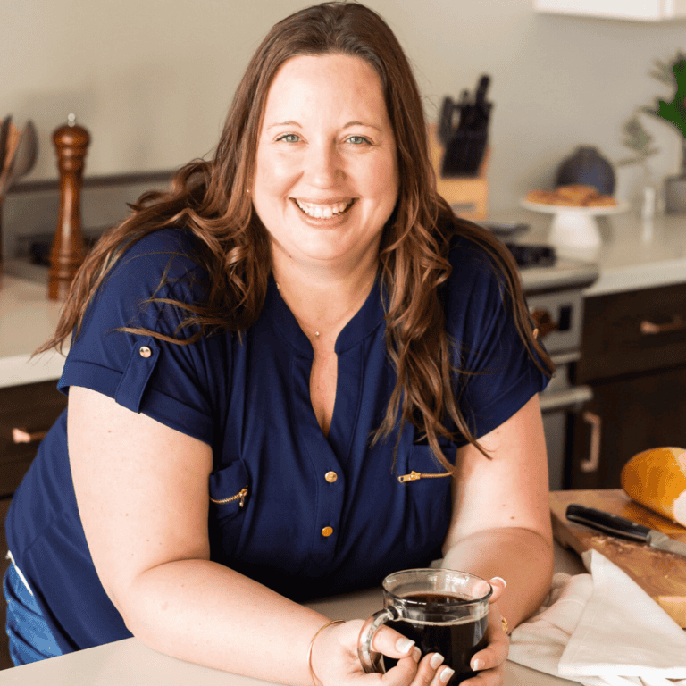 A woman with long brown hair, wearing a blue shirt, is smiling and holding a cup of coffee in a kitchen. There are kitchen utensils and a loaf of bread on the counter in the background.