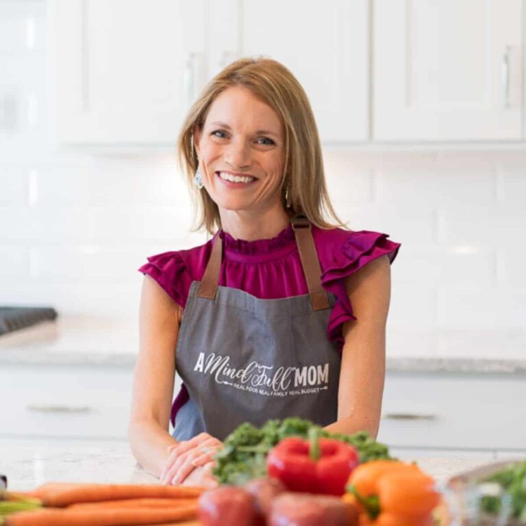 Woman wearing a gray apron with "A Mind 'Full' Mom" written on it, smiling in a kitchen. She's surrounded by vegetables like carrots and peppers, and standing near a counter. The kitchen has a modern white tile backsplash.