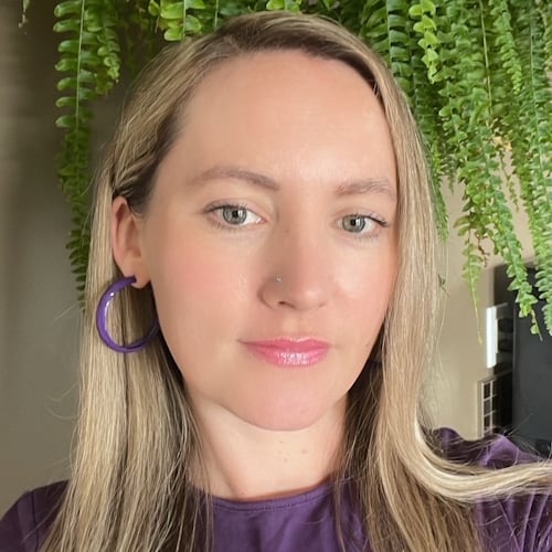 Woman with long blonde hair, wearing a purple top and hoop earrings, standing indoors. A lush green plant hangs behind her, adding a nature-inspired background to the scene.