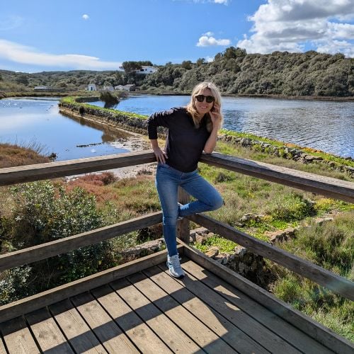 A person wearing sunglasses poses on a wooden platform overlooking a scenic river and lush greenery under a partly cloudy sky. The individual rests one foot on the railing, surrounded by vibrant landscape and a distant house visible in the background.
