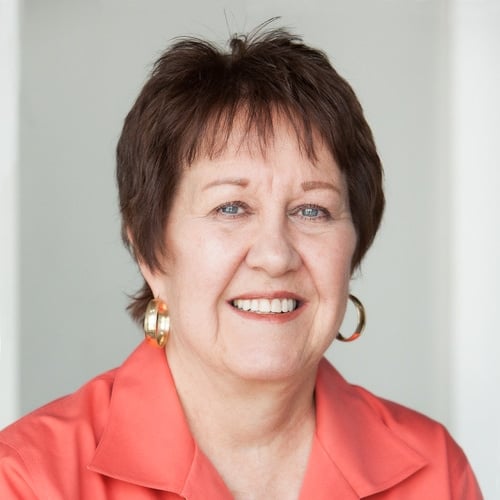 A woman with short brown hair smiles at the camera. She is wearing a coral shirt and gold hoop earrings, set against a plain white background.