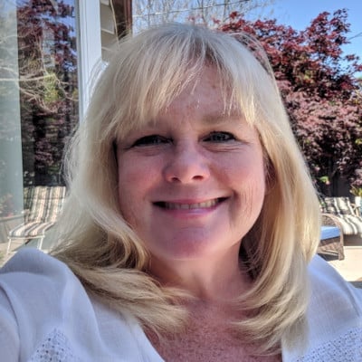 A woman with blonde hair smiles at the camera. She is wearing a white top and is outside, with a view of trees and a patio in the background. The lighting is bright and the atmosphere appears sunny.