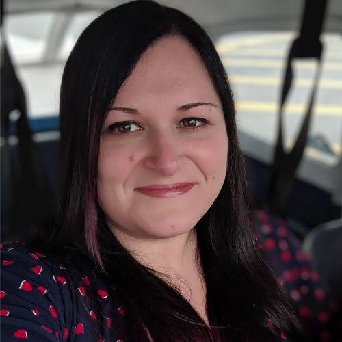 A woman with long black hair and a friendly smile is sitting in what appears to be an airplane. She is wearing a dark-colored top with a pattern of small red hearts. The background shows airplane seatbelts and a window with a glimpse of the tarmac outside.