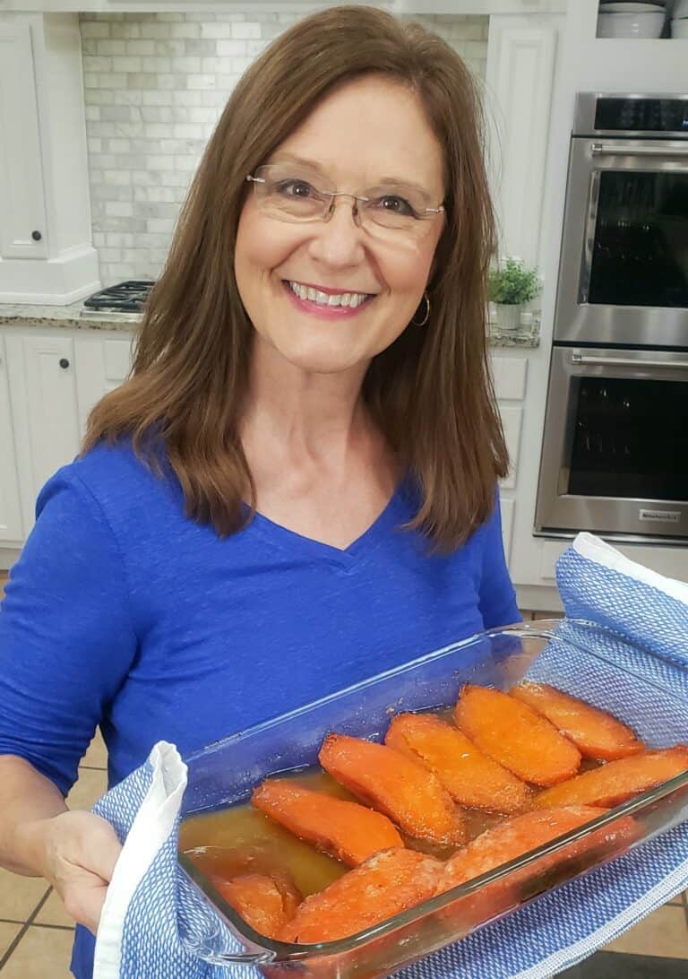 A woman with long brown hair and glasses smiles at the camera while holding a glass baking dish filled with cooked sweet potato wedges. She is wearing a blue shirt and using a blue and white cloth to hold the dish. The kitchen background has white cabinets.