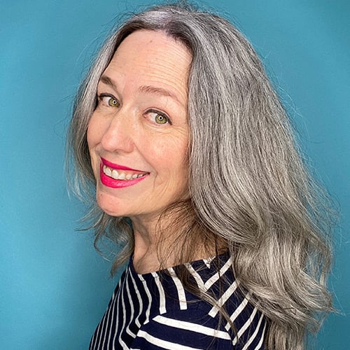 A person with long gray hair smiles at the camera against a blue background. They are wearing a black and white striped shirt and have bright pink lipstick.
