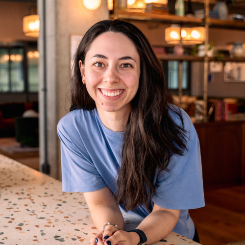 A person with long dark hair and a blue T-shirt is smiling while leaning on a speckled counter. The background features bookshelves with decorations and warm lighting.